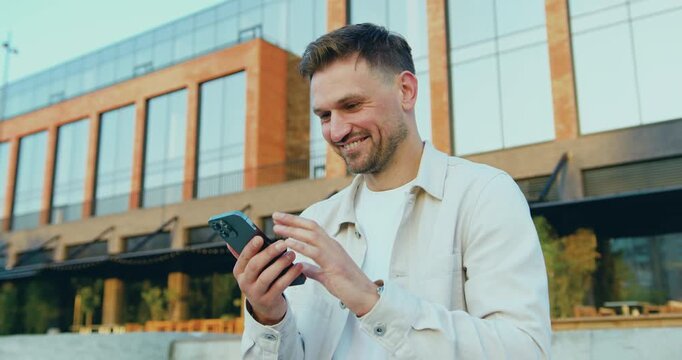 Portrait of a handsome bearded man looking at his smartphone screen in a city park, thoughtfully calculating, outdoors in an urban environment.