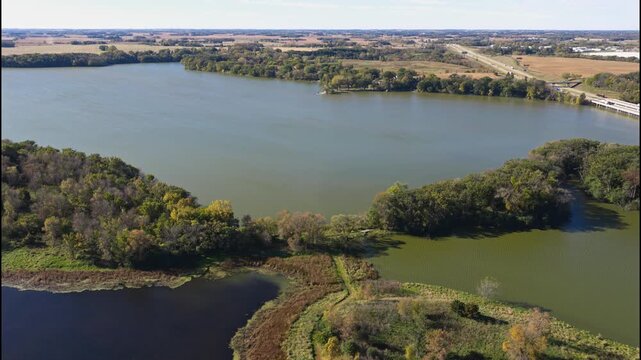 Large lake bordered by autumn woodland and farmland near Albert Lea as cars drive along highway, Minnesota, aerial establishing of Myre-Big Island State Park