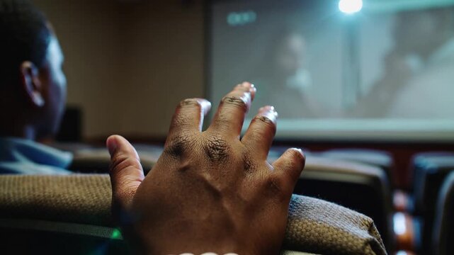 Reacting to projector lamp, man reaching right hand over seat in screening room, with bead bracelet