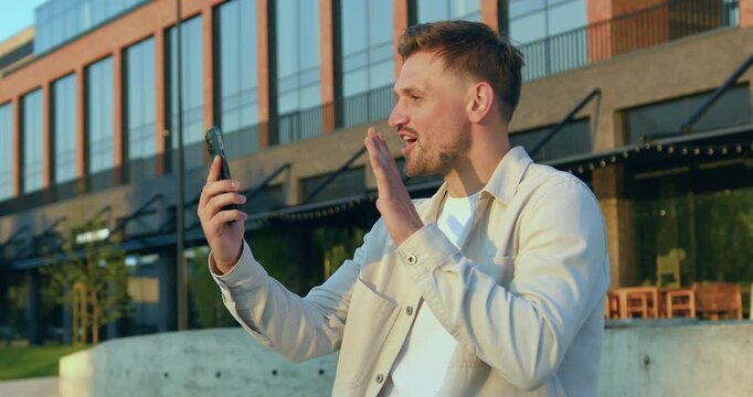 Man stands in front of modern glass office building, holding smartphone in front of him while having an online video call with colleague outdoors.