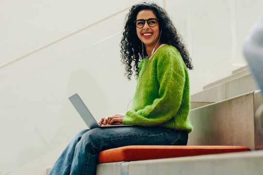 The smiling student studies on a laptop with headphones in library stair seating