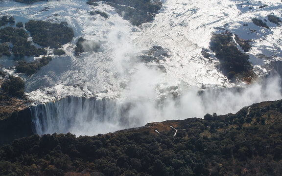 Aerial view of Victoria Falls with water cascading over the edge into the deep gorge, surrounded by lush forest and rising mist in Victoria Falls, Matabeleland North Pr, Zimbabwe.