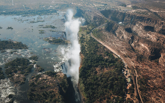 Aerial view of Victoria Falls, showing mist rising from the Zambezi River, lush gorge vegetation, and surrounding arid terrain in Victoria Falls, Matabeleland North Pr, Zimbabwe.