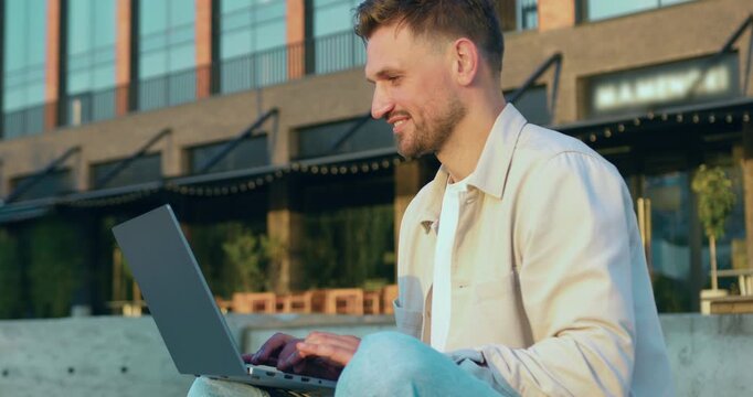 Handsome man entrepreneur using a wireless laptop outdoors near a modern office building, typing and scrolling while managing online business and remote work.