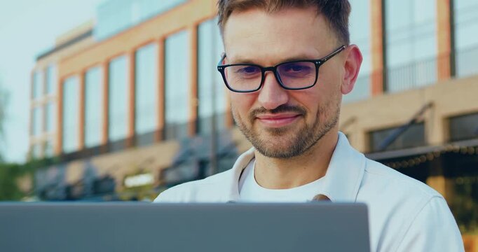 Close up. Portrait of focused Caucasian businessman in a formal suit working remotely on a wireless laptop while standing outdoors in front of a modern glass office building