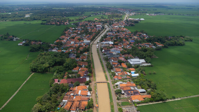 Aerial view of a village with terracotta-roofed houses bisected by a river, surrounded by expansive green rice paddies in Indramayu, West Java, Indonesia.