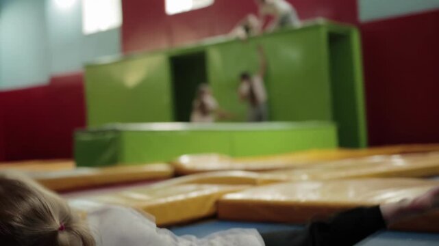 Blonde girl child wearing white t shirt and black pants falls backward onto soft blue mat within spacious indoor play area featuring padded obstacles for children slow mo