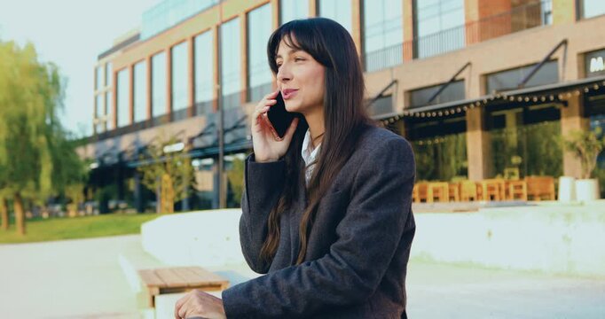 Beautiful young woman making phone call on mobile phone and taking notes while working remotely while sitting on bench about modern office building, outdoors