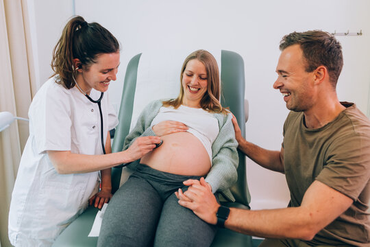 Midwife Listening To Pregnant Belly With Stethoscope As Couple Laugh
