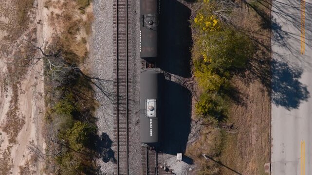 Freight railcars pulled by locomotive near parallel railroad tracks and road in Gentry, Arkansas, aerial top-down tracking