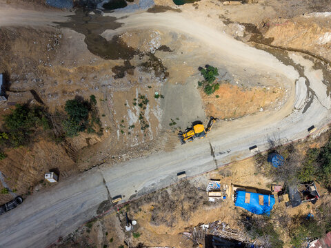 Aerial view of a yellow backhoe loader working on a dirt road at a construction site with surrounding dry terrain and scattered structures in Istanbul, Istanbul, Turkiye.