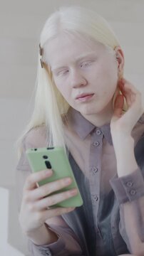 Stylish albino lady using smartphone focused on internet communication sitting on stairs in luxury apartment. Creative interior and online content concept.