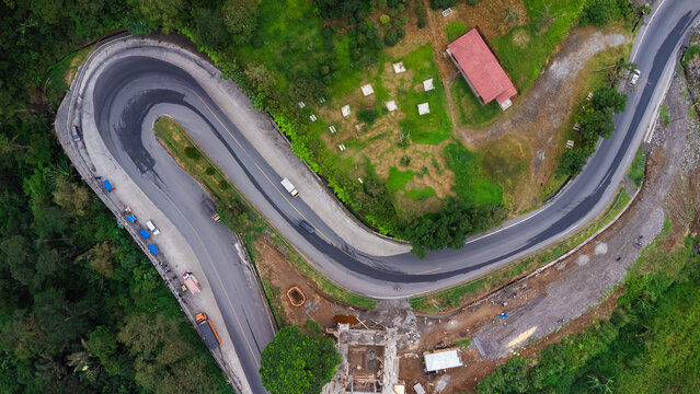 Aerial view of the winding road at Penatapan Berastagi with sharp hairpin turns surrounded by lush tropical forest and construction areas in Sibolangit, North Sumatra, Indonesia.