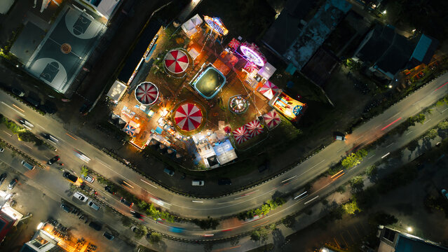 Aerial view of a vibrant night carnival with illuminated rides and colorful tents surrounded by city roads with light trails from moving vehicles in Depok, West Java, Indonesia.
