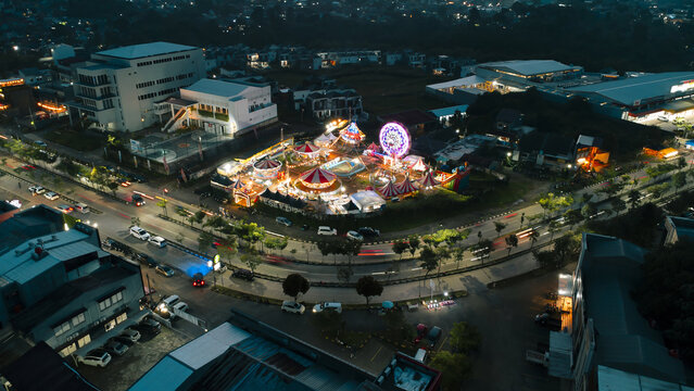 Aerial view of a vibrant night carnival with a glowing ferris wheel and illuminated tents situated next to a busy road with light trails in Depok, West Java, Indonesia.