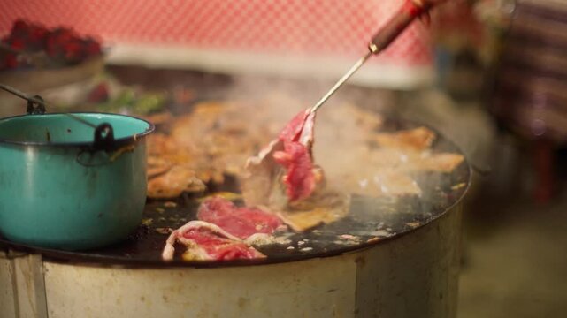 Meat cooks on a hot griddle beside a steaming pot during community celebration, spatula flips and turns beef slices