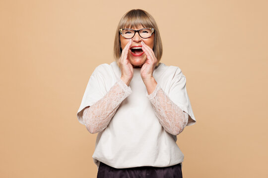 Elderly blonde woman 50s years old wear white shirt casual clothes scream sharing hot news about sales discount with hand near mouth isolated on plain pastel light beige background. Lifestyle concept