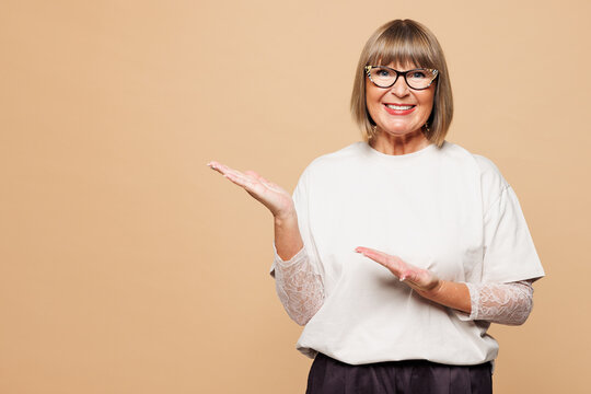 Elderly blonde woman 50s years old she wears white shirt casual clothes point hands arms aside on area look camera isolated on plain pastel light beige background studio portrait. Lifestyle concept.