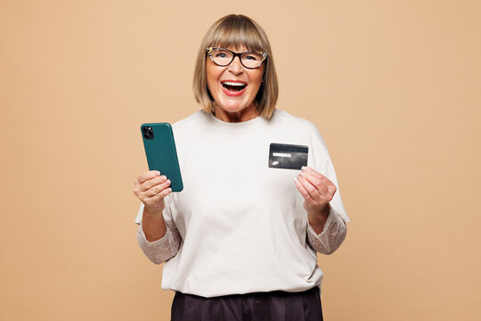 Elderly woman 50s years old wear white shirt casual clothes using mobile cell phone credit bank card shopping online order delivery booking tour isolated on plain beige background. Lifestyle concept.