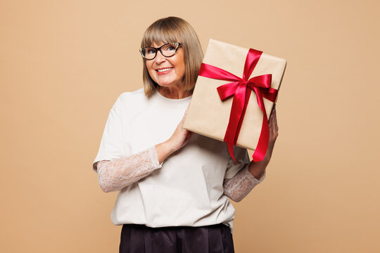 Elderly blonde fun woman 50 years old she wearing white shirt casual clothes hold present box with gift ribbon bow look aside isolated on plain pastel light beige background studio. Lifestyle concept