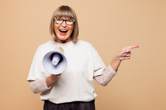 Elderly blonde woman 50s years old wear white shirt casual clothes hold megaphone scream announces discounts Hurry up point finger aside on area isolated on plain beige background. Lifestyle concept.