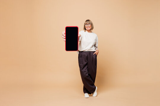 Full body elderly blonde woman 50s years old she wear white shirt casual clothes hold use show blank screen mobile cell phone isolated on plain pastel light beige background studio. Lifestyle concept.