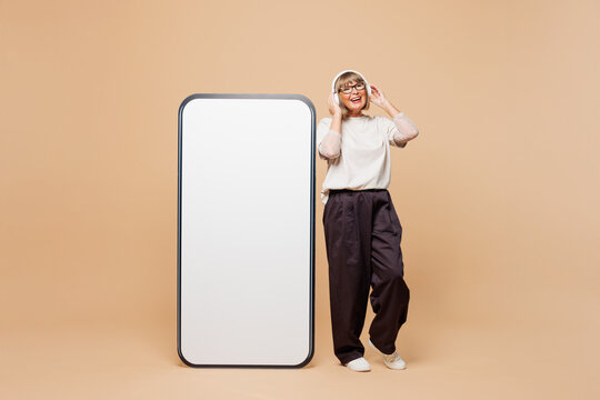 Full body elderly woman 50 years old wear white shirt casual clothes big huge blank screen mobile cell phone with area listen music in headphones isolated on plain beige background. Lifestyle concept