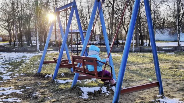 Young child in winter clothing plays on swing at playground during golden hour. Sun shines through bare trees, illuminating patches of snow on ground.