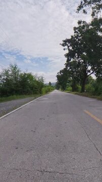 Vertical hyperlapse traveling along a winding rural road with guardrails and chevron signs, surrounded by green vegetation under a cloudy blue sky.