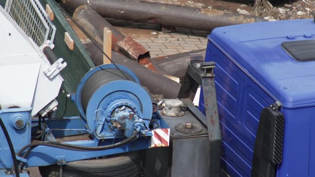 Close-up view of blue winch mechanism with wire rope on heavy construction truck crane, showing industrial equipment details and discarded metal pipes in background
