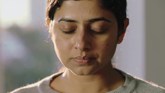 Closing eyes, woman sensing sunlight and making subtle mouth motion at window, grey sweater