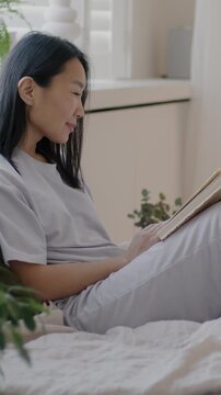 Slow motion of carefree Asian lady in pajamas sitting in bed reading book smiling relaxing in morning in luxurious bedroom at home. Literature and apartment concept.