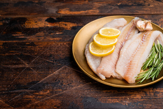 Pollock sea fish fillets, raw whitefish on a plate with herbs and lemon. wooden background. top view