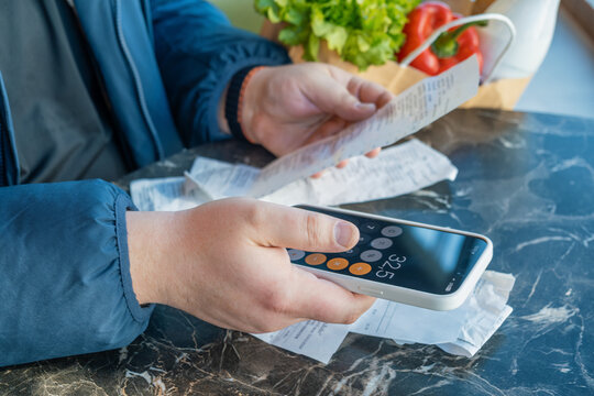 Shopper uses smartphone calculator while reviewing grocery receipts beside bag of everyday food. Rising shopping expenses, inflation, cost of living pressure, household financial budgeting