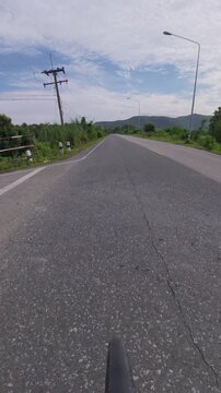 Vertical POV footage of a cyclist riding along a scenic rural road toward distant mountains, surrounded by lush greenery and roadside street lights under a cloudy blue sky.