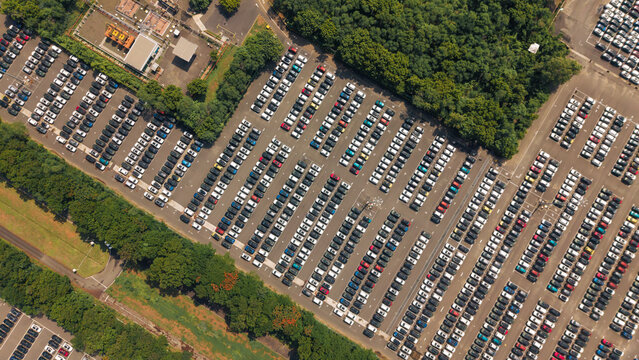 Aerial view of a car manufacturing unit parking lot filled with rows of new vehicles, surrounded by lush green trees and industrial infrastructure in India.