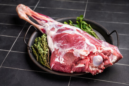 Uncooked whole goat leg, raw meat with herbs for cooking. black background. top view