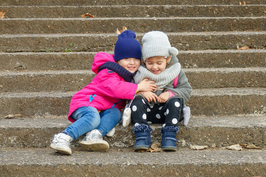 Two Happy Girls Hugging and Laughing Together on Outdoor Stone Steps in Warm Winter Clothes, Friendship and Childhood Joy