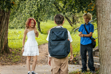 Obraz premium Caucasian children playing outdoors in a park, one girl in a white dress holding a stick, two boys with backpacks engaging in conversation on a sunny day