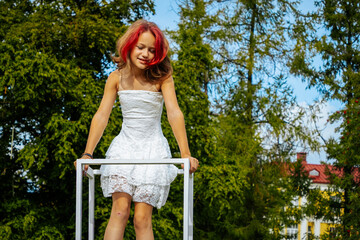 Young girl with red hair wearing a white dress stands on a white structure outdoors, surrounded by green trees and a building in the background under a blue sky © Jolanta