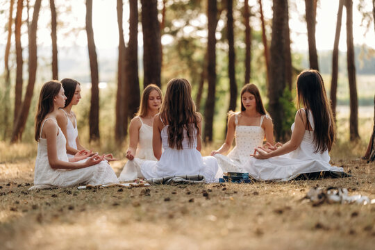 Group of young women in white dresses meditating together on grass under trees in a serene outdoor setting during daylight with soft sunlight filtering through branches