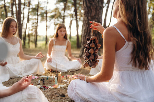 Group of young women in white dresses participating in a spiritual ritual, sitting in a circle on the ground surrounded by natural scenery and decorative elements