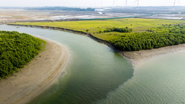Aerial view of a winding river flowing through lush green mangroves and coastal fields with wind turbines in the distance in Moheskhali Upazila, Chittagong Division, Bangladesh.