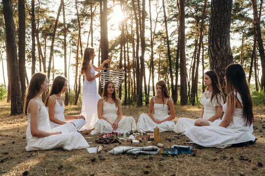 Group of women in white dresses sitting in a circle on the ground during a gathering in a forest, with sunlight filtering through the trees in the background