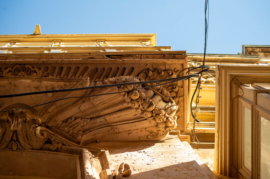 Low angle close-up of sculpted limestone architectural details on a historic building exterior. Intricate stone corbel and relief carving are highlighted in warm sunlight under a clear sky.