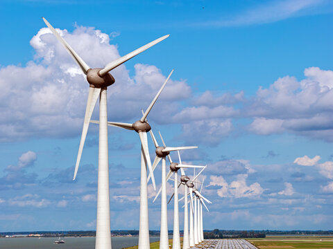 Aerial view of a row of wind turbines standing in the flat landscape near the water under a blue sky with clouds in Rutten, Flevoland, Netherlands.