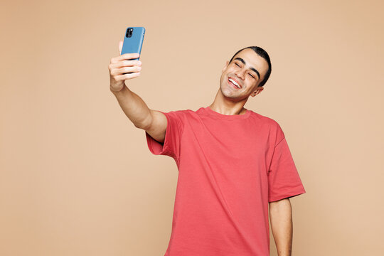 Young middle eastern man wear red t-shirt casual clothes doing selfie shot on mobile cell phone post photo on social network isolated on plain pastel light beige background studio. Lifestyle concept.