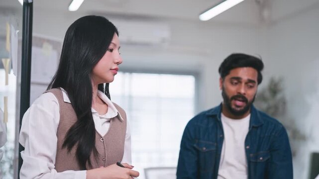 Creative businesswoman brainstorming and writing project strategy on a clear glass board with a marker while her colleague watches carefully.