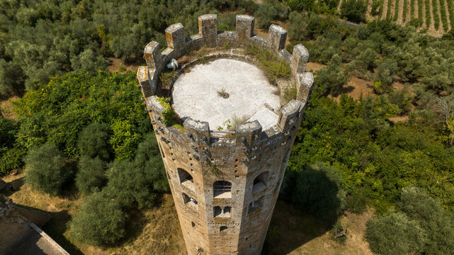 Aerial view of an ancient crenellated tower amid olive groves in the Italian countryside. The circular stone tower features arched window openings and a flat rooftop terrace.