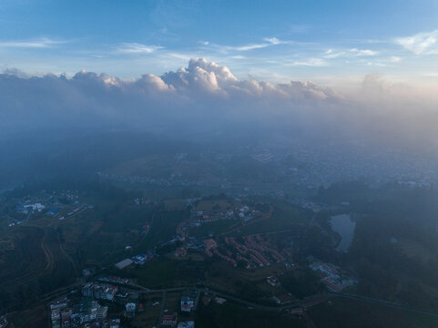 Aerial view of Ooty nestled among rolling hills with scattered buildings, a small lake, and a blanket of mist under a soft sky in Ooty, Tamil Nadu, India.